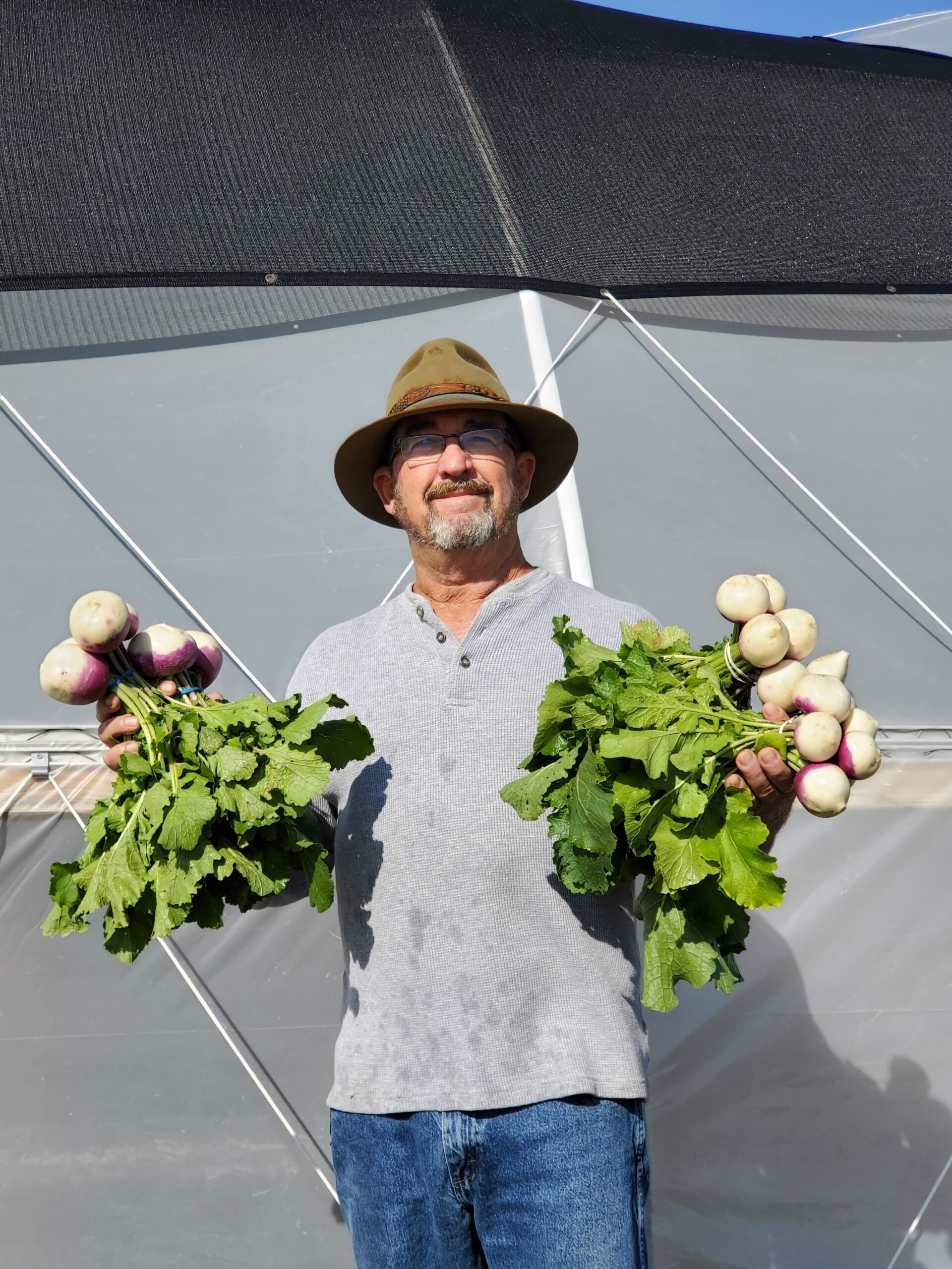 A man wearing a hat smiles while holding two bunches of freshly harvested turnips and radishes in front of a greenhouse.