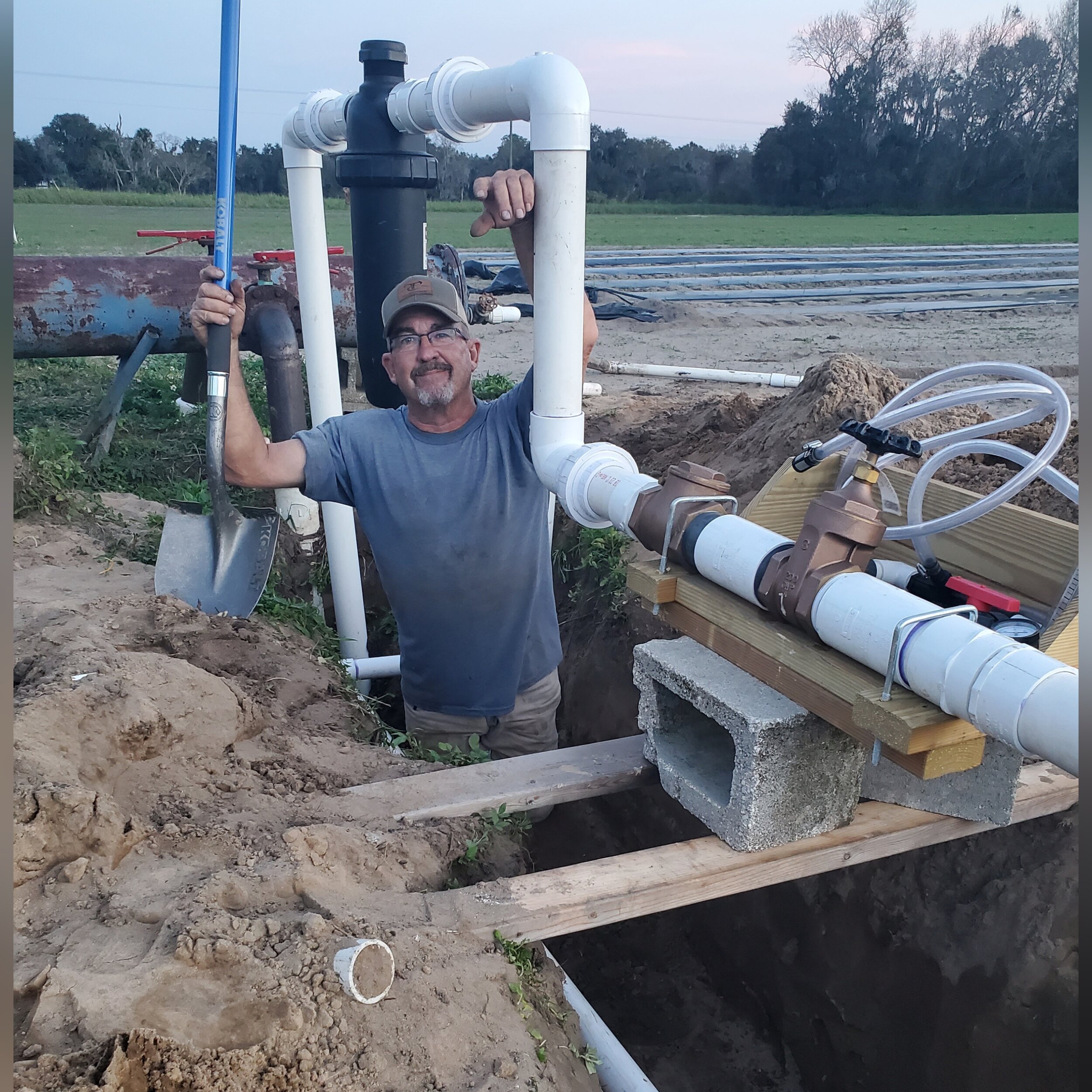 Man standing in a trench with plumbing pipes, holding a shovel and smiling, surrounded by agricultural land and equipment. The image showcases the installation of irrigation systems with PVC pipes and valves.
