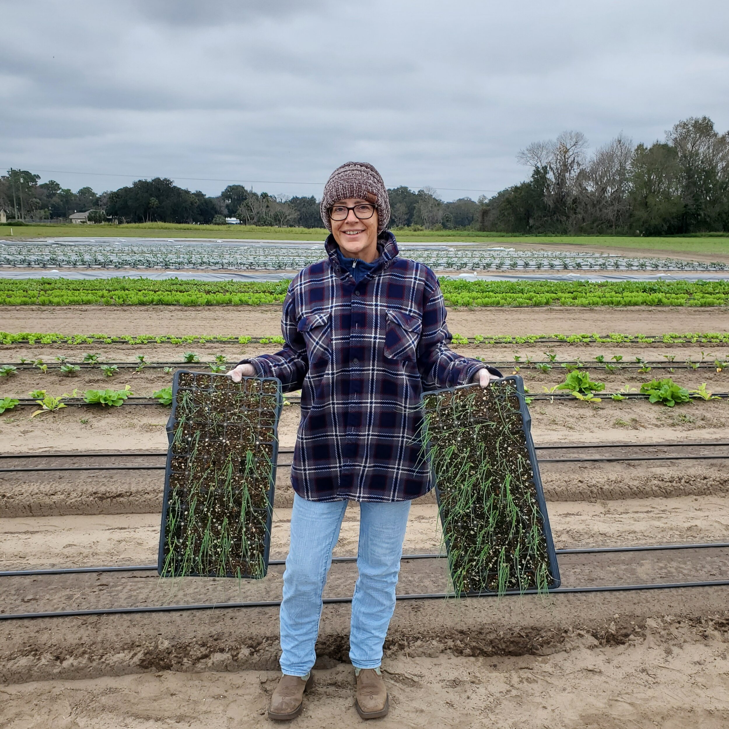 Woman in a plaid shirt and knit hat holding seedlings in trays on a farm, with rows of lettuce and other crops in the background.