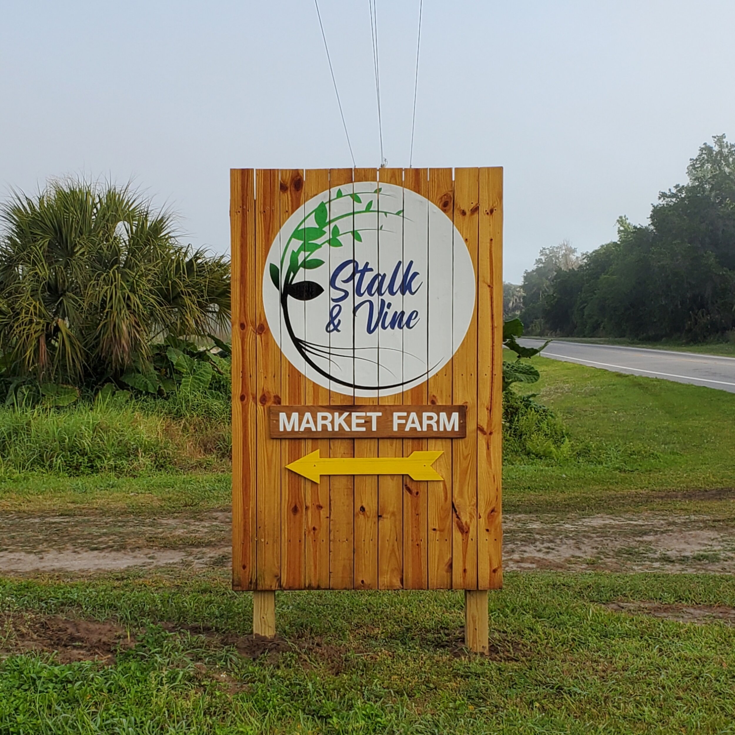 Wooden sign for Stalk & Vine Market Farm, featuring a circular logo with green plant graphics, located near a roadside with lush greenery in the background. The sign directs visitors to the market farm with an arrow pointing left.
