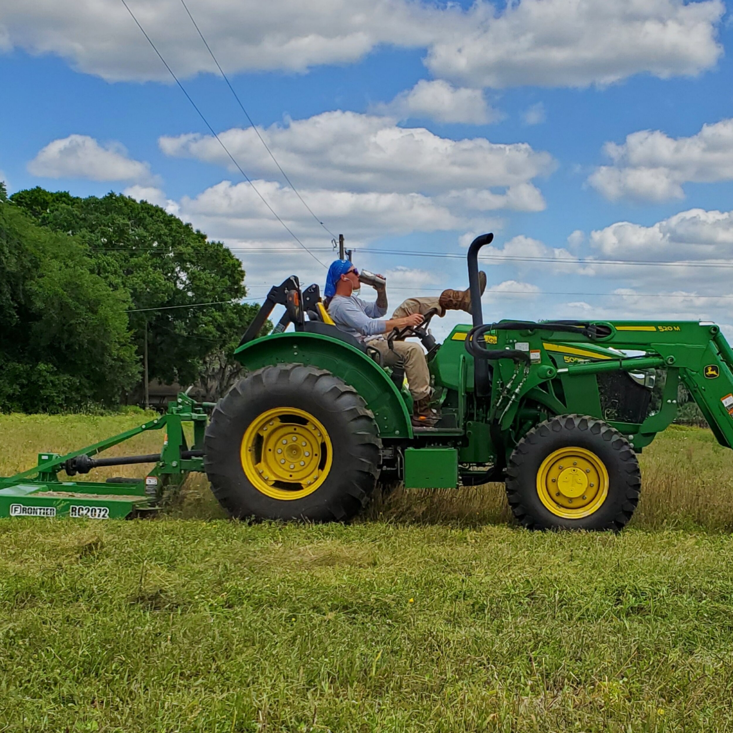 A farmer operating a green John Deere tractor equipped with a Frontier RC2072 mower, cutting grass in a sunny field under a blue sky with fluffy clouds.
