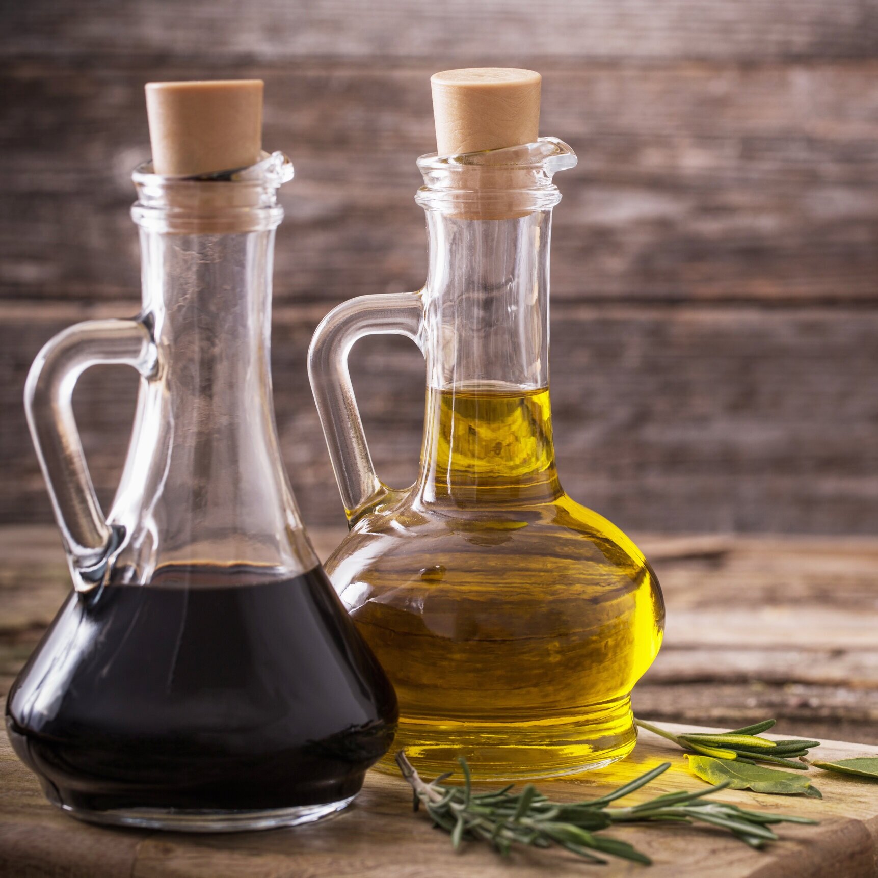 Two glass bottles of olive oil and balsamic vinegar with cork stoppers, placed on a wooden surface alongside fresh rosemary sprigs.